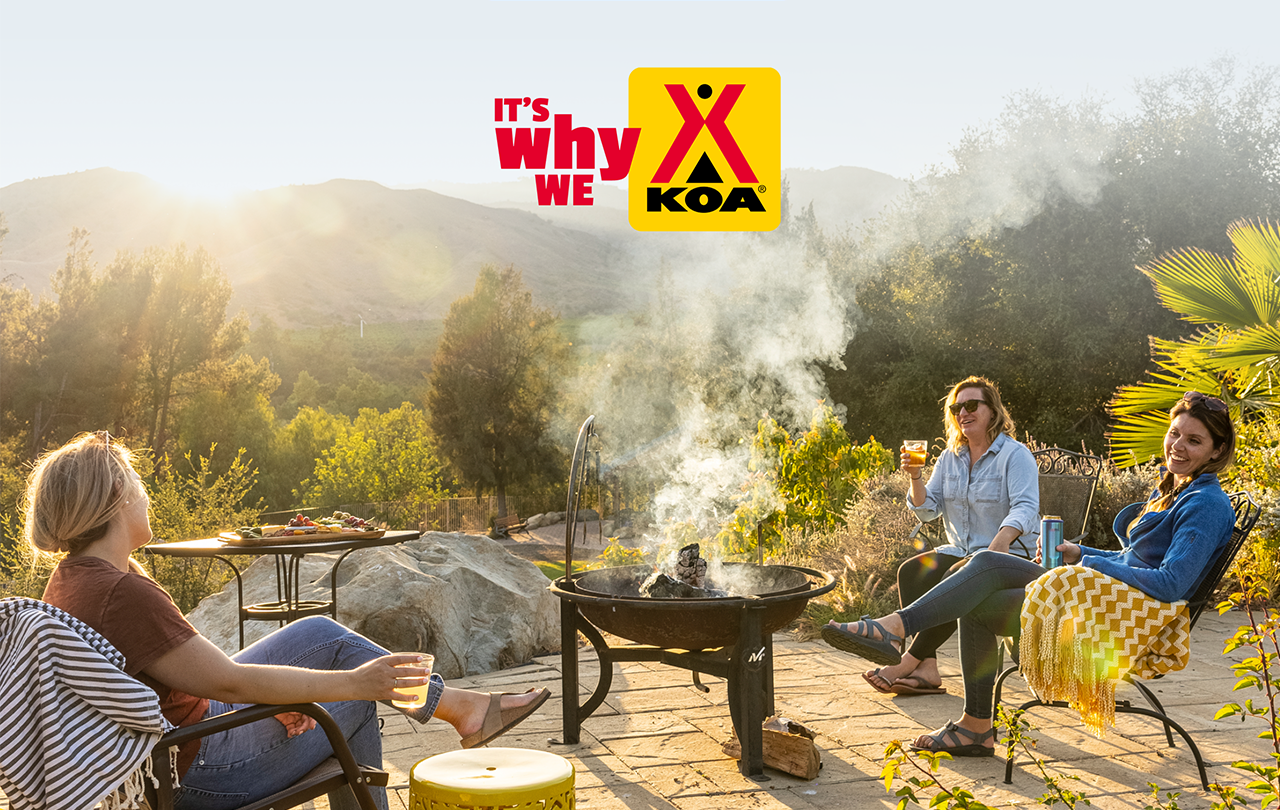 Three women sitting around a firepit with mountains in the distance. A logo says It's Why We KOA.