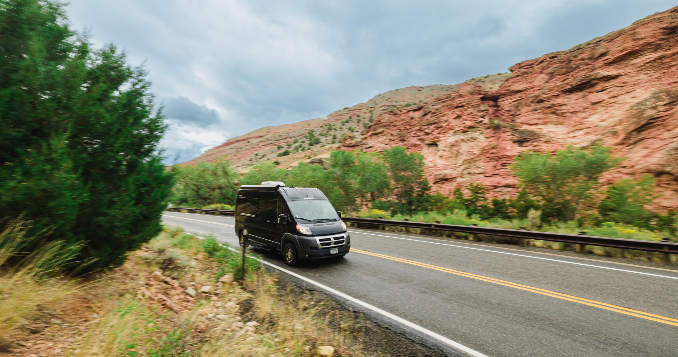 A black campervan driving down a road with rocky bluffs in the distance.