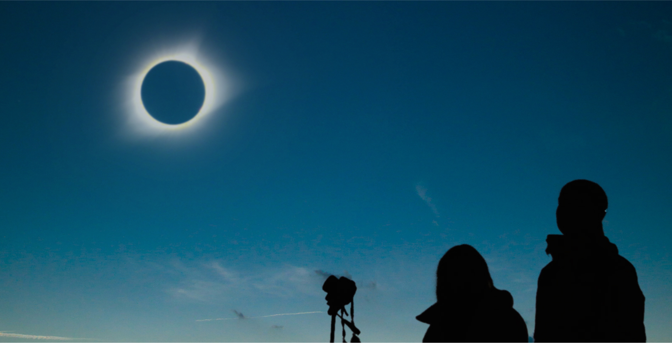 Silhouettes of two people and a camera looking at a total solar eclipse in the sky.