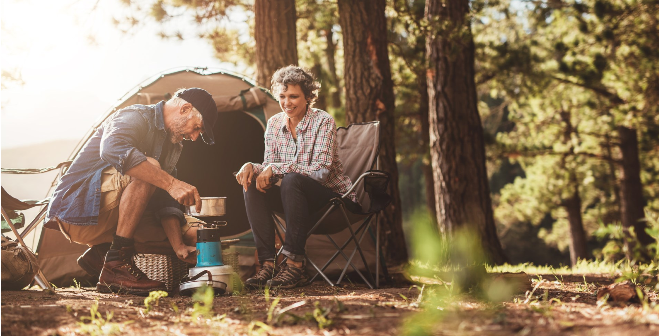 A middle aged couple cooking outside their tent in the woods.