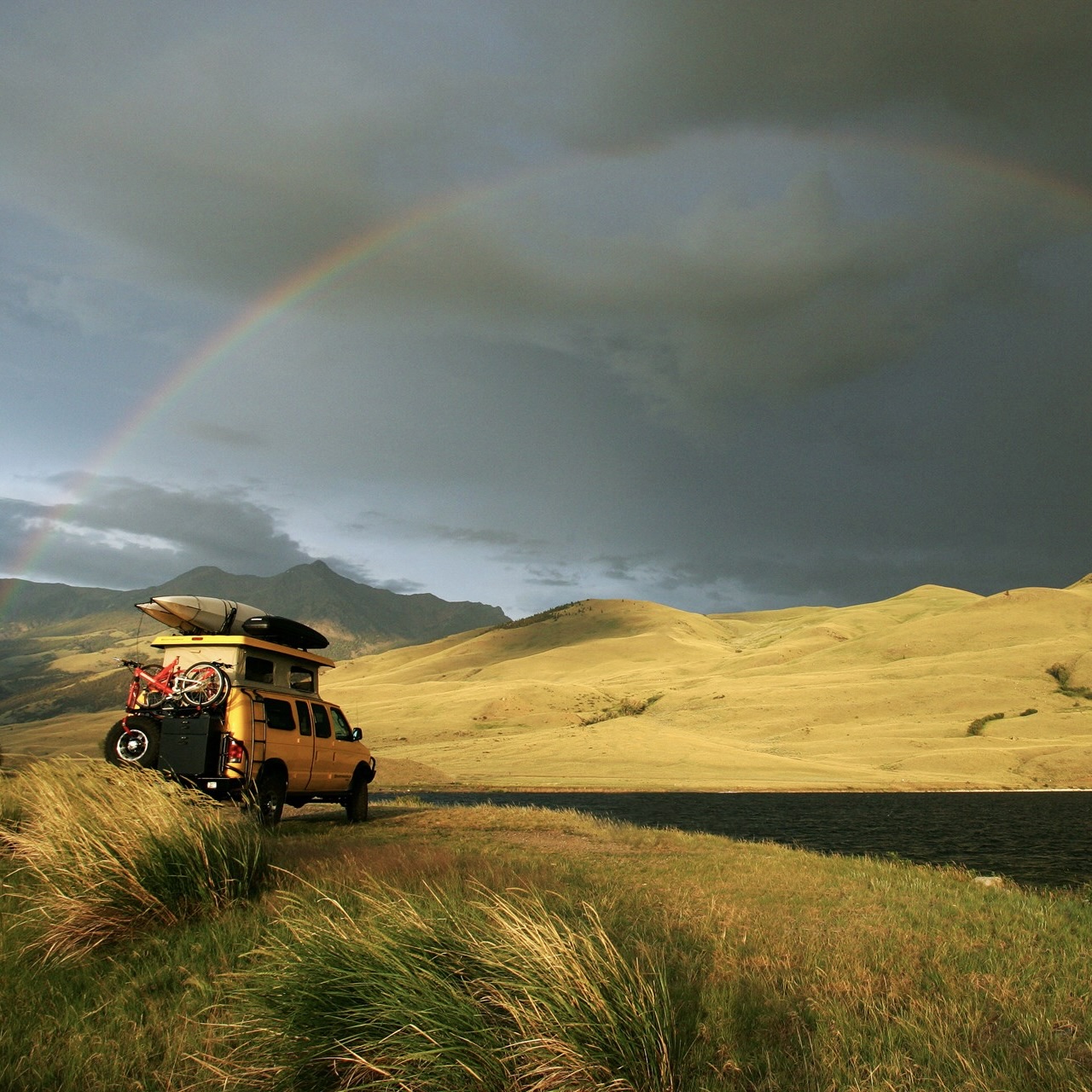 A yellow van parked in a field under a cloudy sky and a rainbow