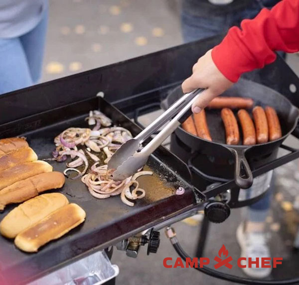 Food being cooked on an outdoor griddle at a tailgate party in the daytime.