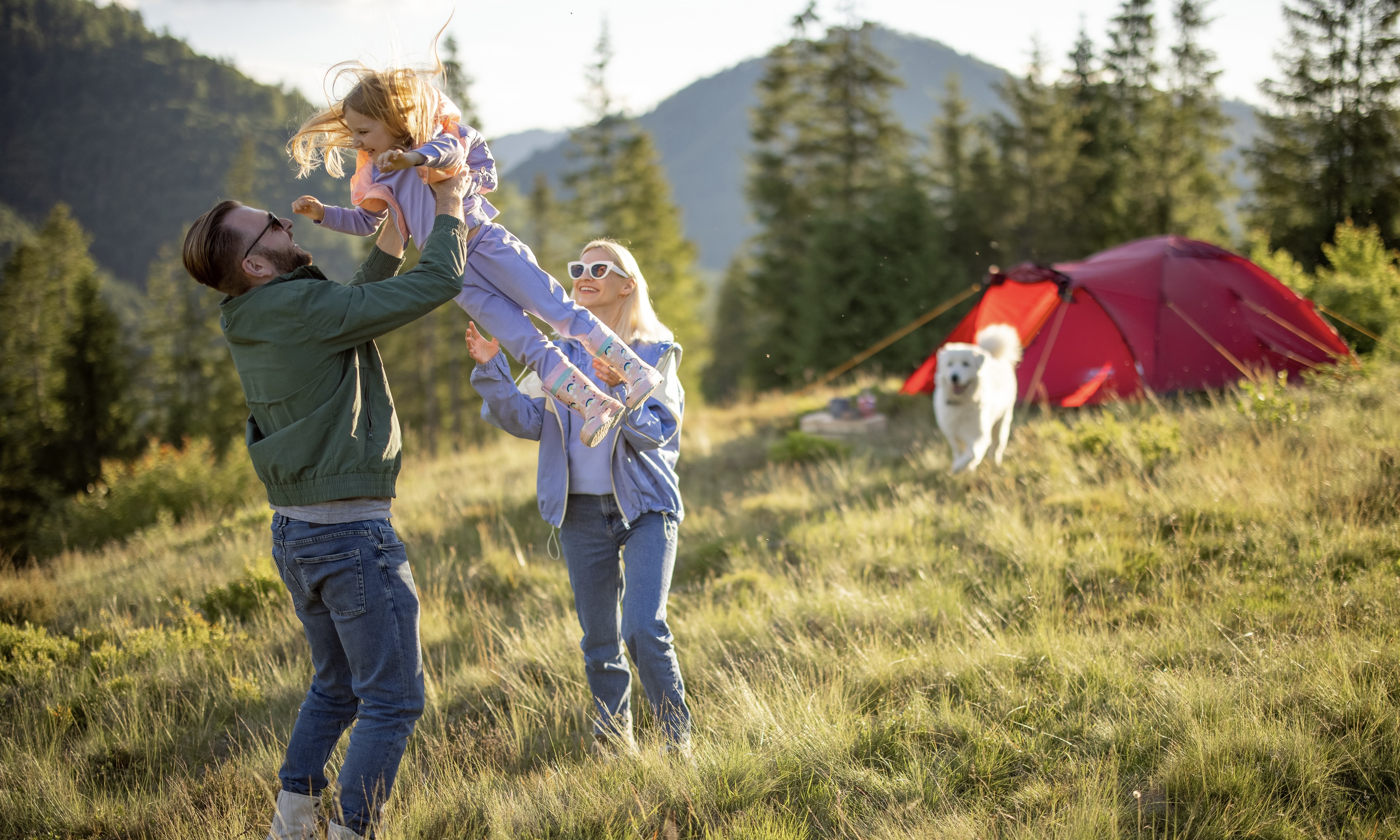 Two parents holding their young daughter at a campsite with a dog in the background